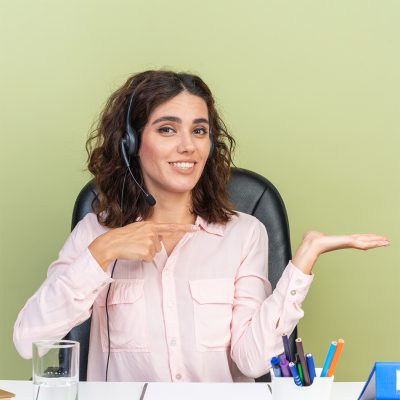 smiling pretty caucasian female call center operator on headphones sitting at desk with office tools pointing at her empty hand isolated on green background